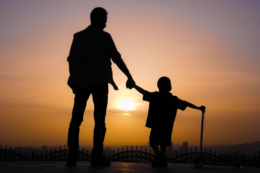 A father and child stand together, holding hands, in silhouette at sunset.