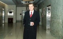 Matt Gaetz, wearing a red tie under a black overcoat, smiles for a camera while standing in front of an elevator lobby.