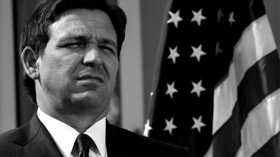 Black-and-white photo of Florida Governor Ron DeSantis squinting, standing in front of an American flag