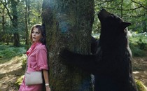 A film still of Cocaine Bear standing on one side of a tree, and a woman hiding on the other