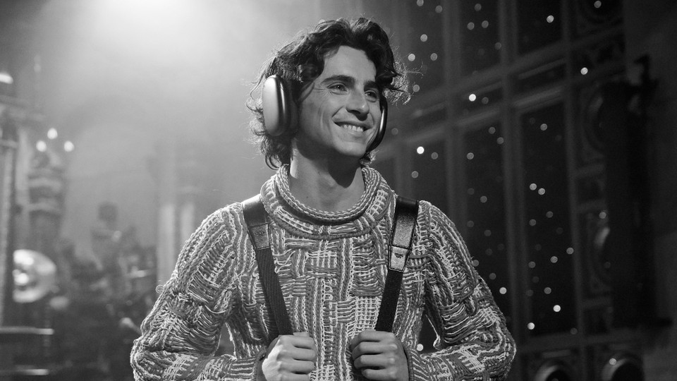 Timothée Chalamet wearing a thick knit sweater holding backpack straps and wearing over-ear headphones, in black and white