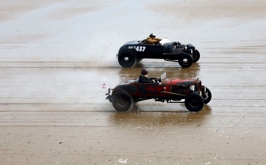 Two people drive old race cars on a flat beach.