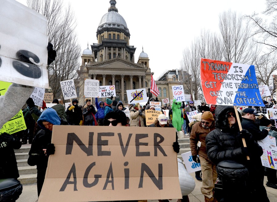 A group of protesters stands on the steps of a state capitol building, holding signs, including one that reads 