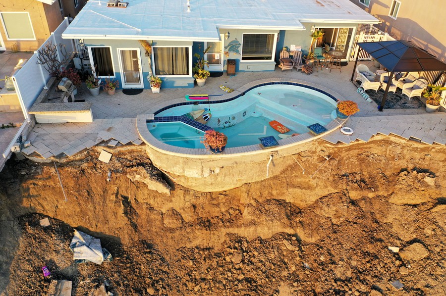 An aerial view of a swimming pool at the edge of a landslide that has cut away much of the surrounding patio.