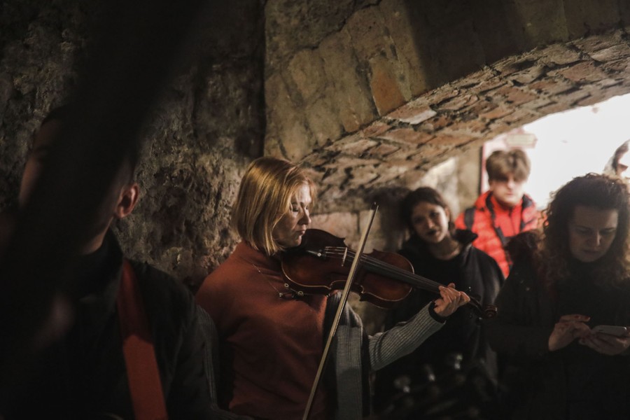 A person plays a violin for others inside a dark shelter with stone walls.