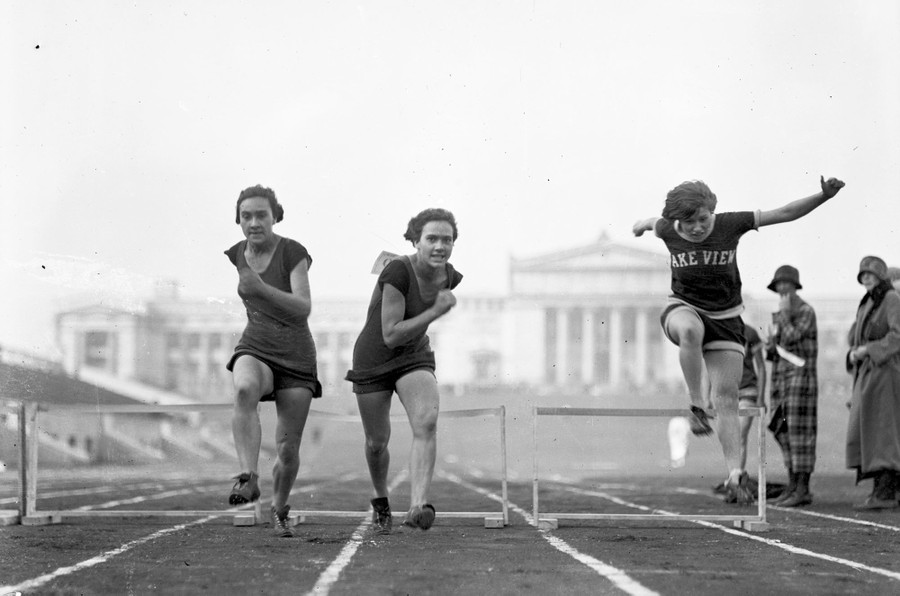 Three runners jump over hurdles on a race track.