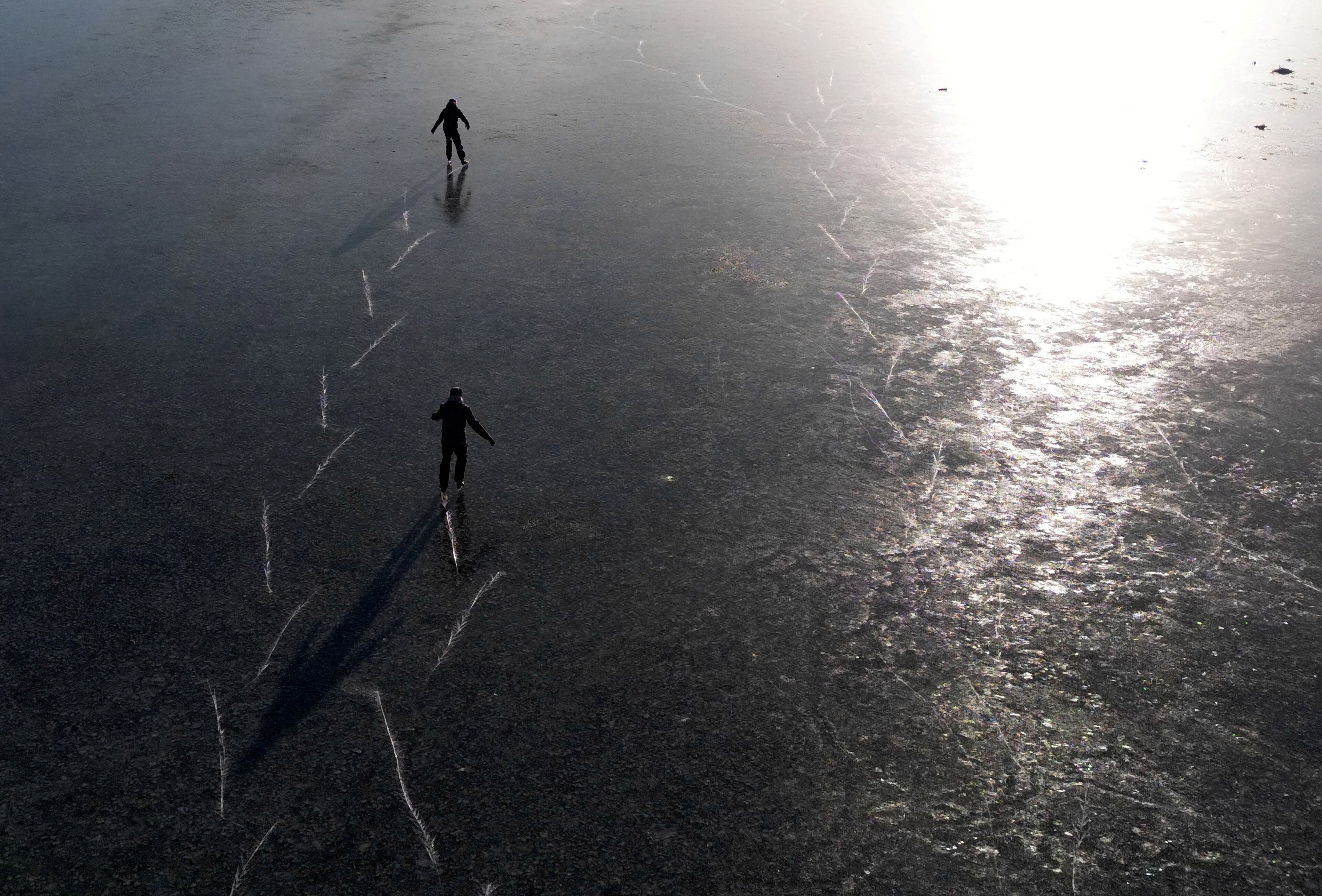 Two skaters leave tracks in ice as they skate over a frozen flooded plain.