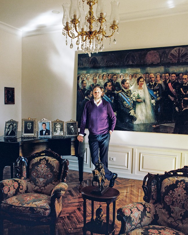 photo of a man in purple sweater standing in front of a large painting of a royal wedding and beside a piano on which five portrait photos are displayed