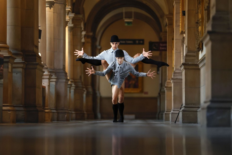 Two dancers in costume pose together, each lifting one leg high, inside a gallery.