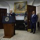 Joe Biden gestures at a White House podium, Miguel Cardona stands by the open door