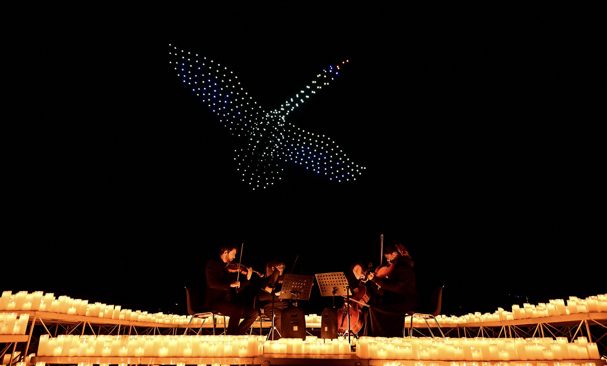 A string quartet performs as drones form the figure of a swan in the night sky above.