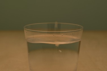 A short glass of water, resting on a light brown table, with an olive green wall in the background.