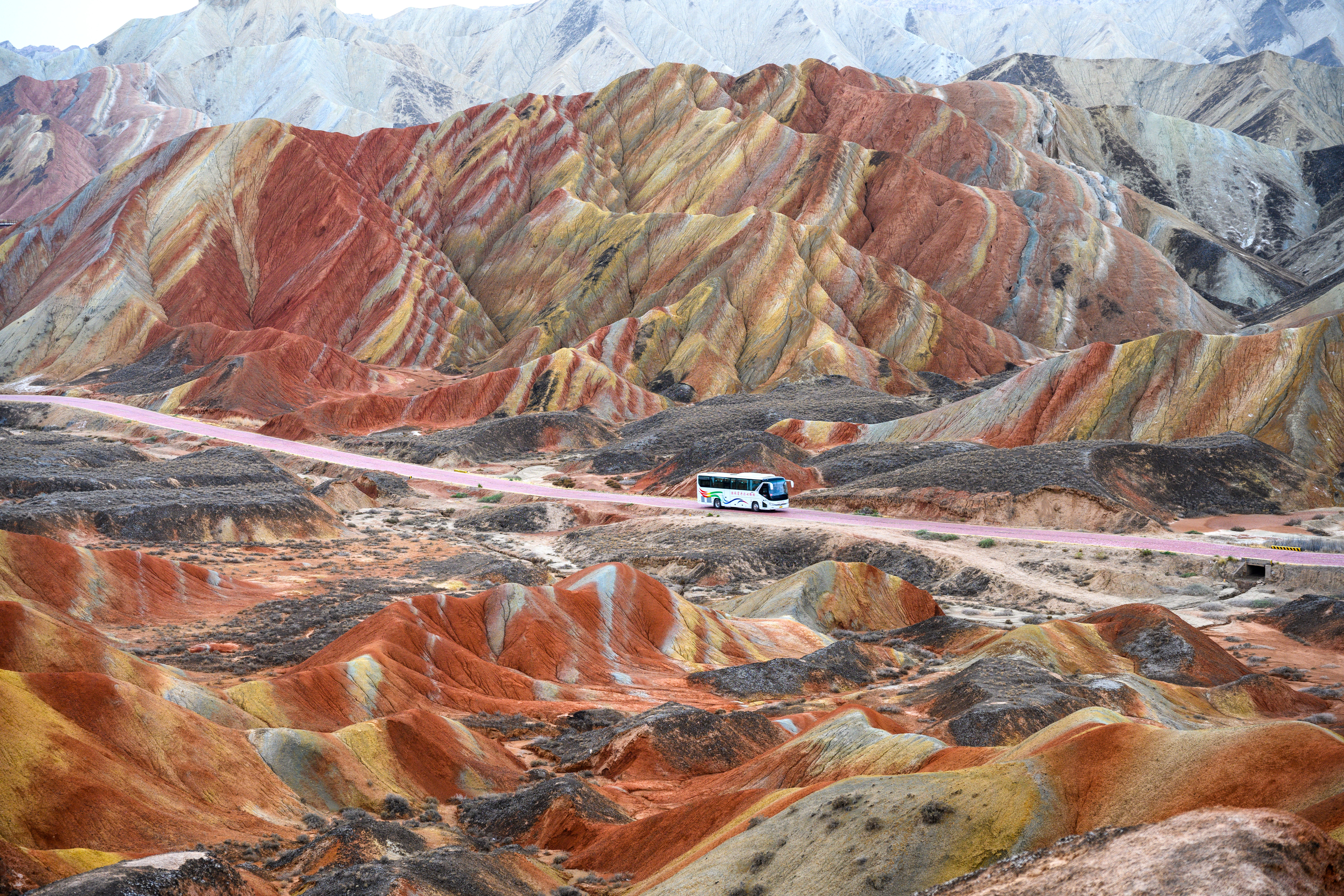 A bus drives down a dirt road through a hilly desert area. The hills are remarkably striped with multiple colors.