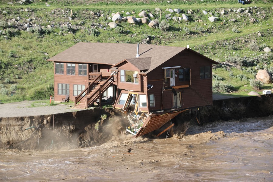 A fast-flowing river undercuts its banks, causing a two-story wooden cabin to start collapsing.