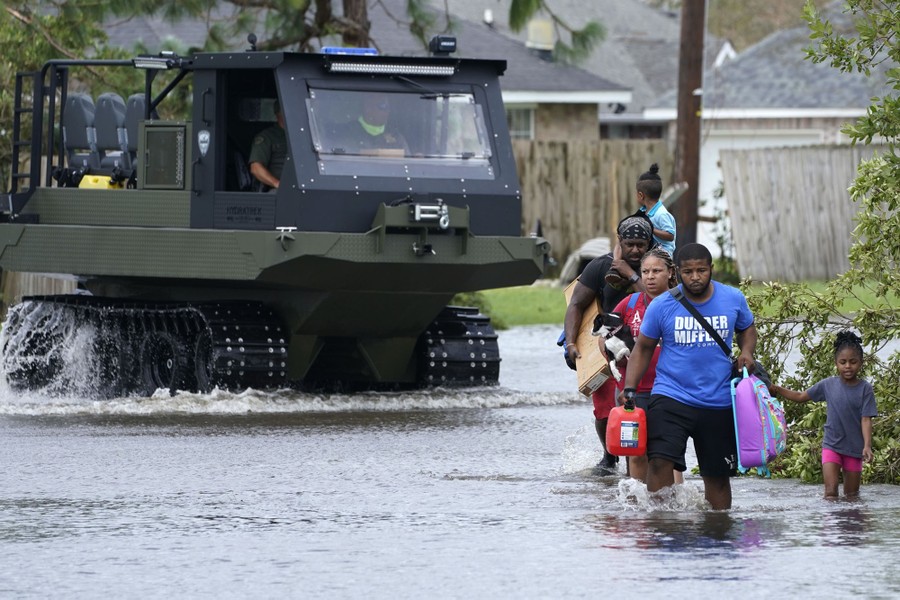 Several adults carry belongings, a pet, and a child in knee-deep flood water, with a rescue vehicle in the background.