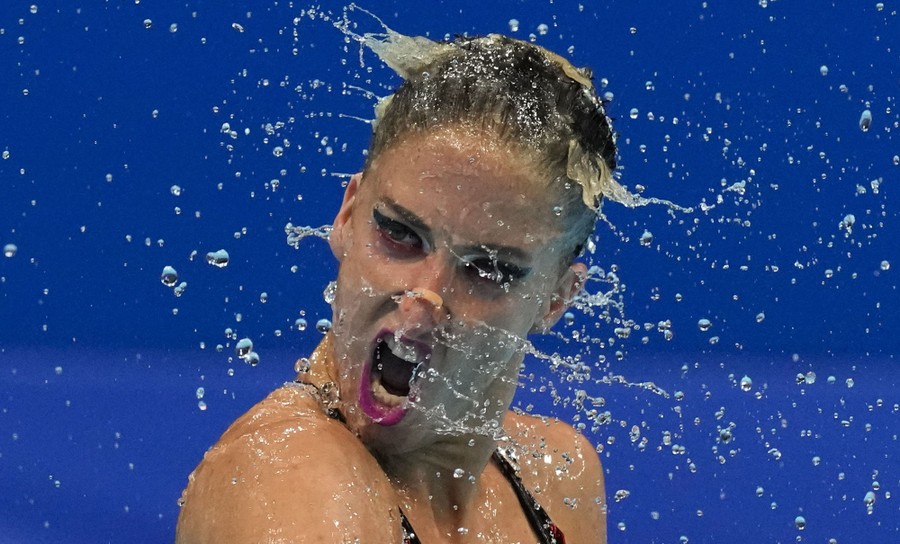 An artistic swimmer twists her head, splashing drops of water away from her.