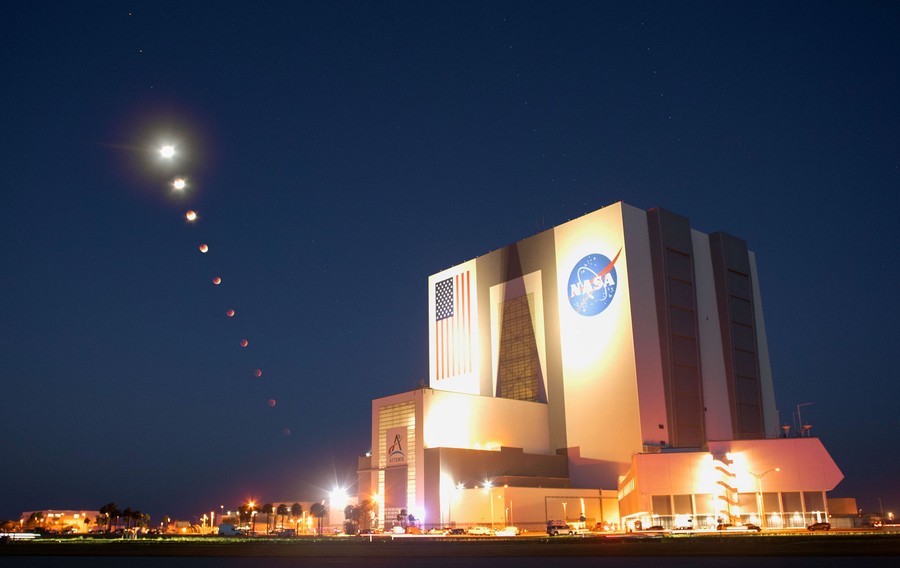 A composite image, showing 10 images of the moon as it drops toward the horizon, dimming with each step, in front of a large NASA building.