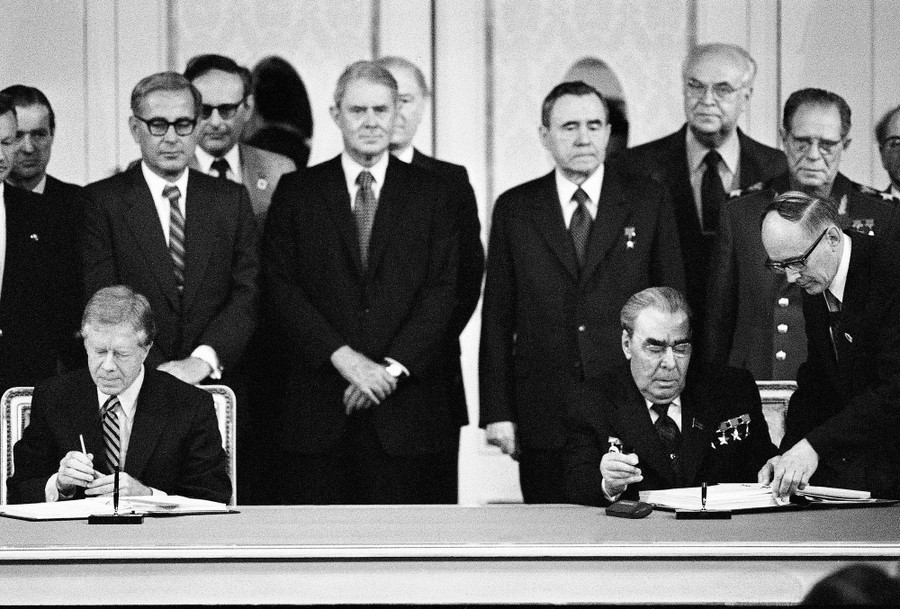 Jimmy Carter and Leonid Brezhnev sign documents in front of a dozen or so officials watching from behind.
