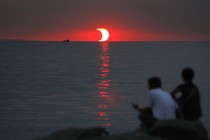 Two people sit on rocks overlooking water, with a partly eclipsed sun setting in the distance.