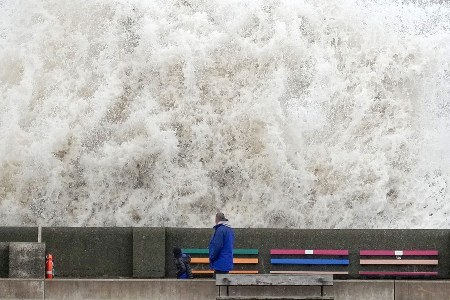 A person looks up at a rising wall of water after a wave crashes into a nearby sea wall.