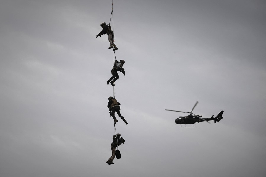 Four soldiers holding weapons are suspended from a single cable beneath an unseen helicopter.