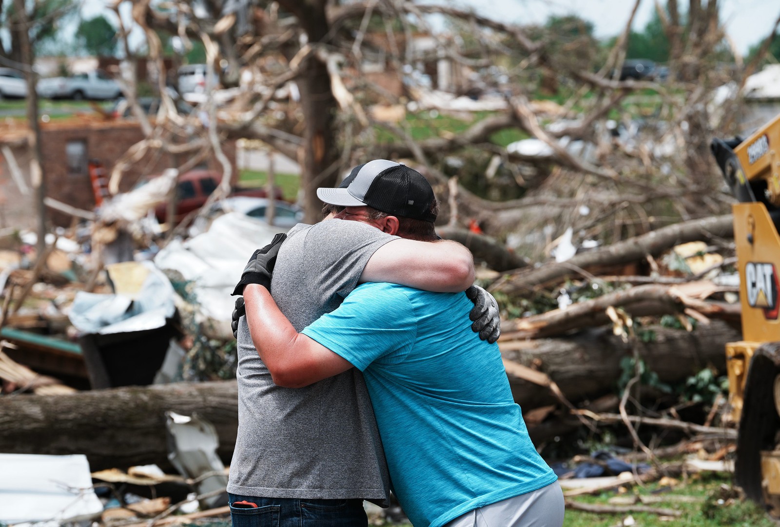 Two people embrace while standing among debris and storm-damaged houses.