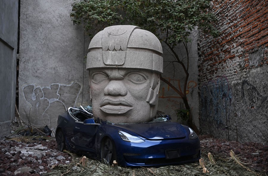 A giant head sculpture sits atop a crushed car in a small lot surrounded by brick and concrete walls.