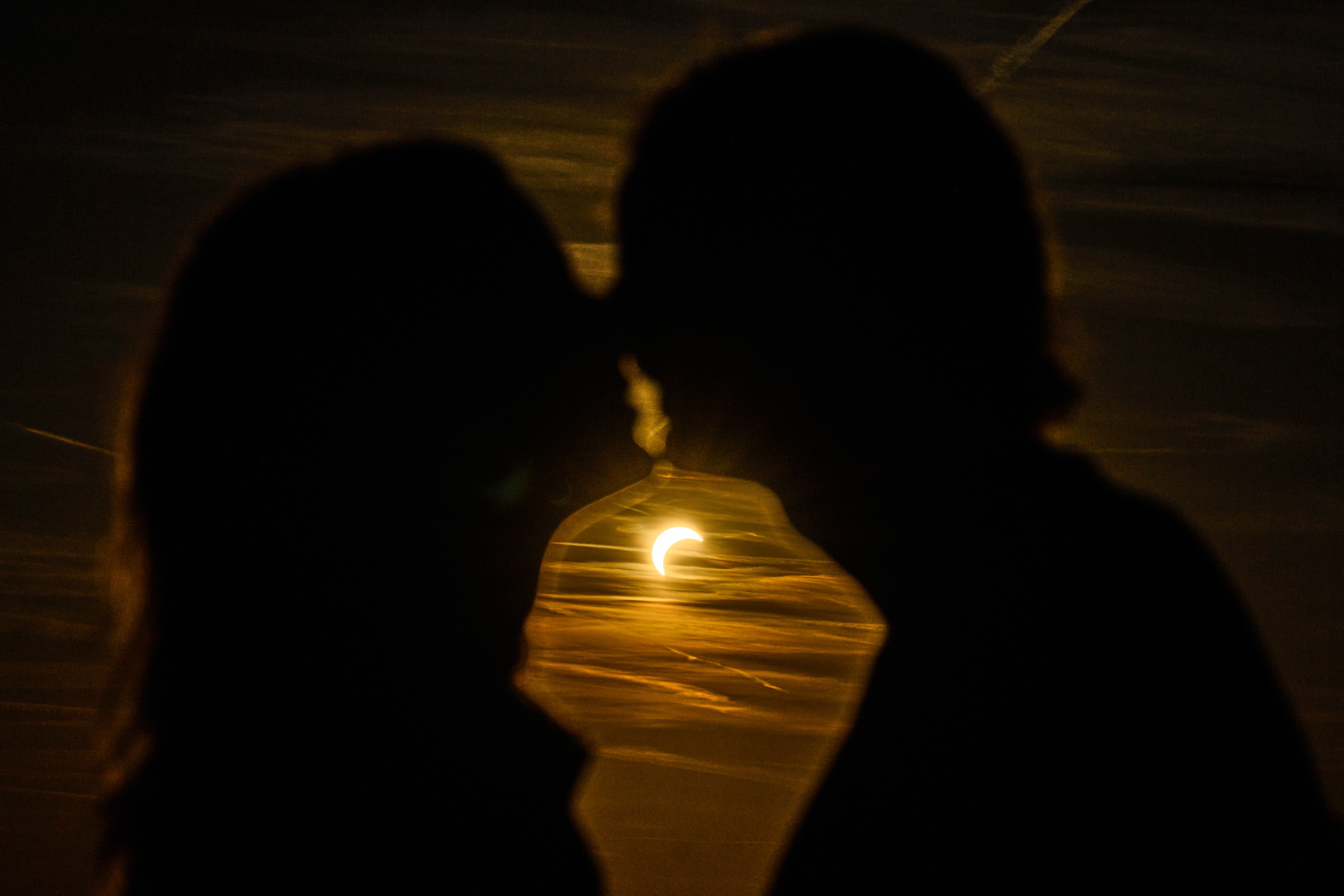 A couple shares a kiss, seen in silhouette, with a partially-eclipsed sun in the background.