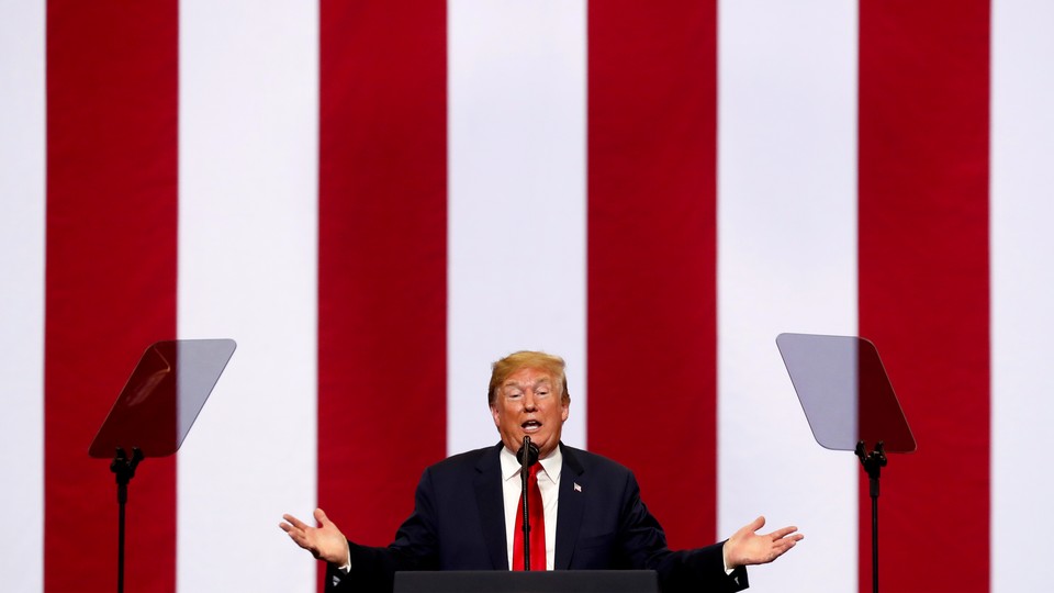 A photograph of Donald Trump standing behind a microphone and a pair of Teleprompters, in front of red-and-white-striped background