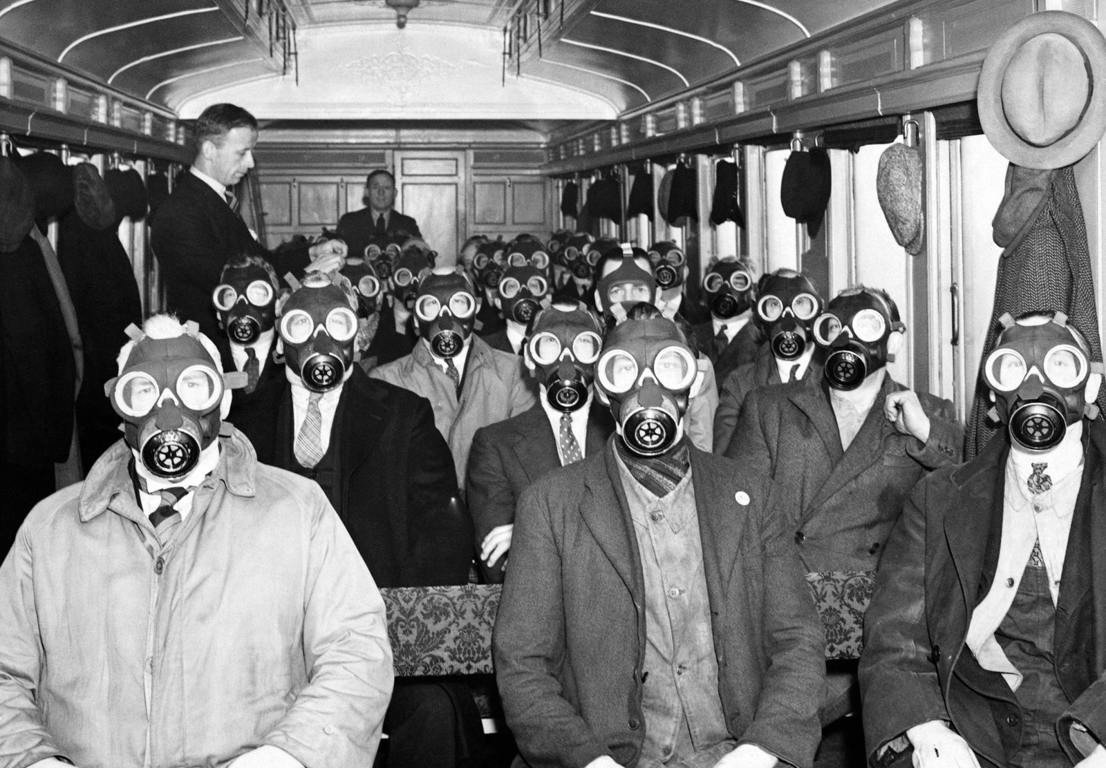 A couple of dozen men sit on benches in a railway car, all wearing gas masks, and facing the camera.