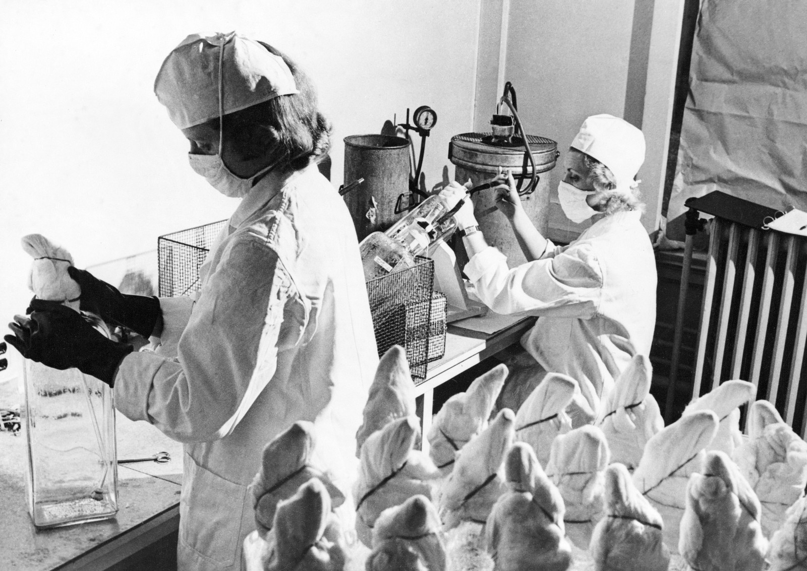 Two medical workers wearing masks, caps, and lab coats work with various equipment to produce vaccines.