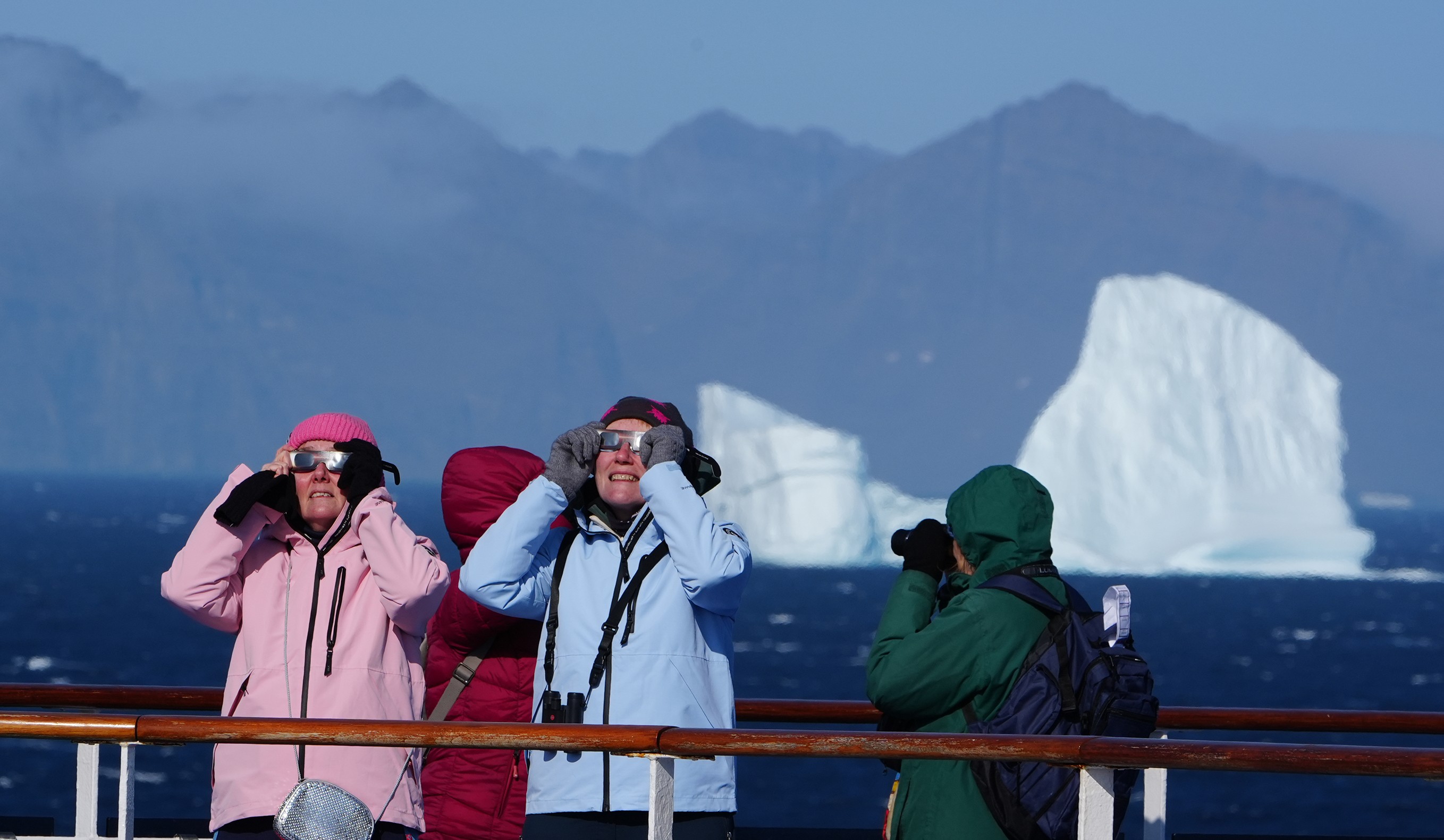 Passengers on board a cruise ship look up into the sky with eclipse glasses on, with icebergs in the background.