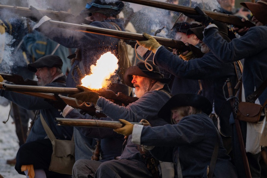 Costumed reenactors line up and fire muskets in a field.
