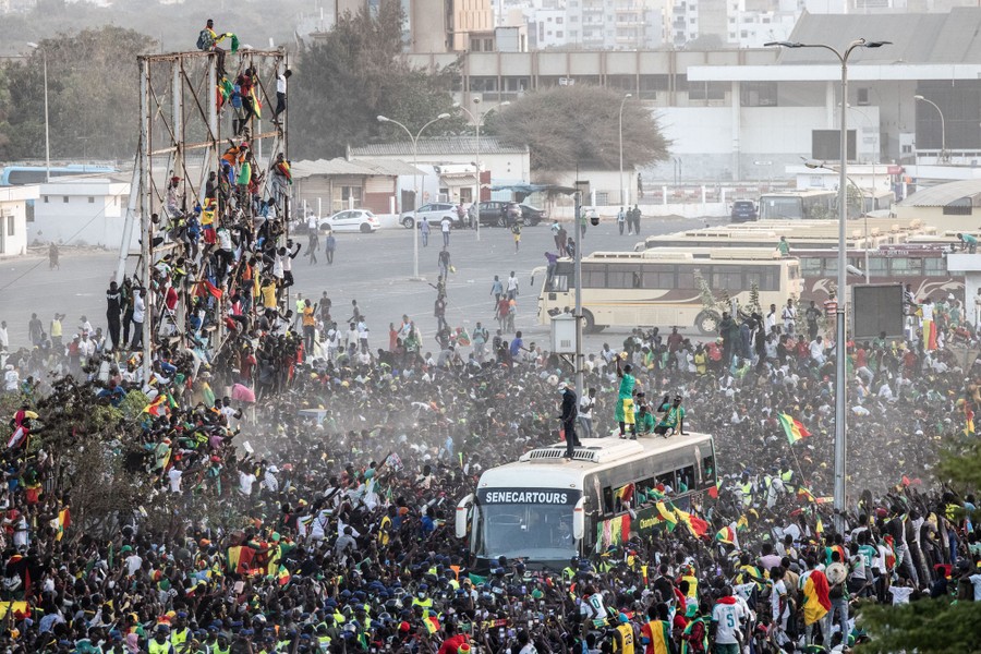 A large crowd of people surrounds a bus on a street, cheering for an athlete who stands on top of the bus.