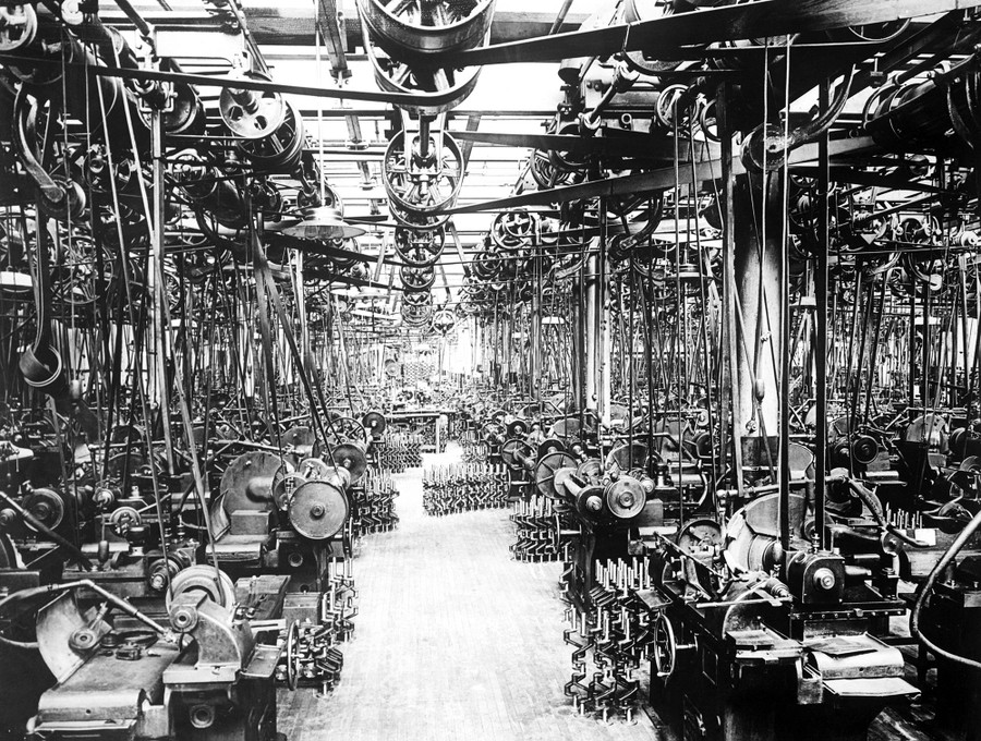 An automobile factory room filled with machinery and drive belts, with stacks of newly built crankshafts on the floor