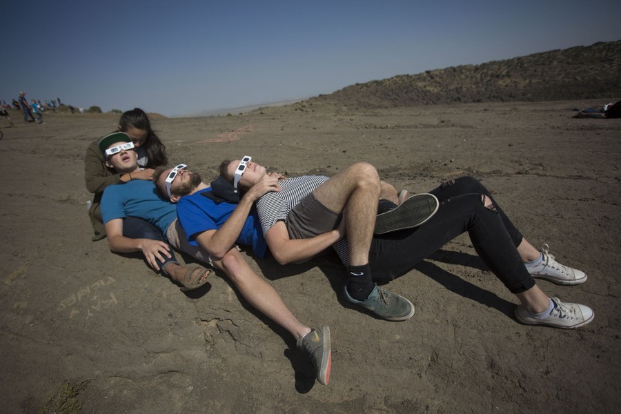 A small group of people lay across each other's laps on rocky ground, wearing eclipse glasses, looking up.