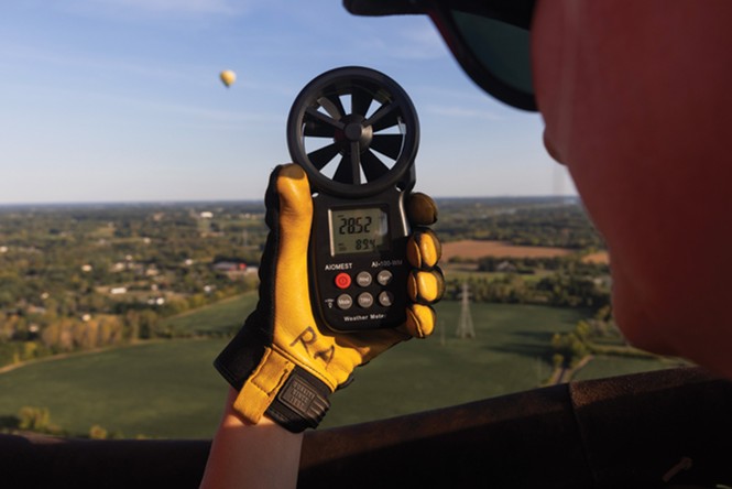 photo of author looking at instrument she holds with gloved hand while flying in balloon, with another hot-air balloon flying in the distance