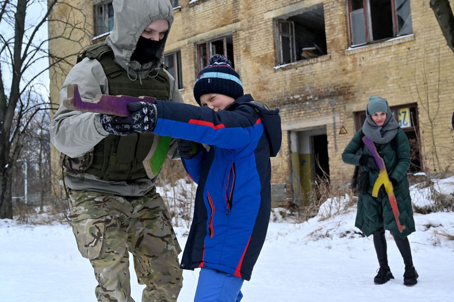 A soldier helps a young person who is holding a wooden replica of a rifle.