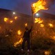 A group of people stand on a hillside, holding torches on long poles.