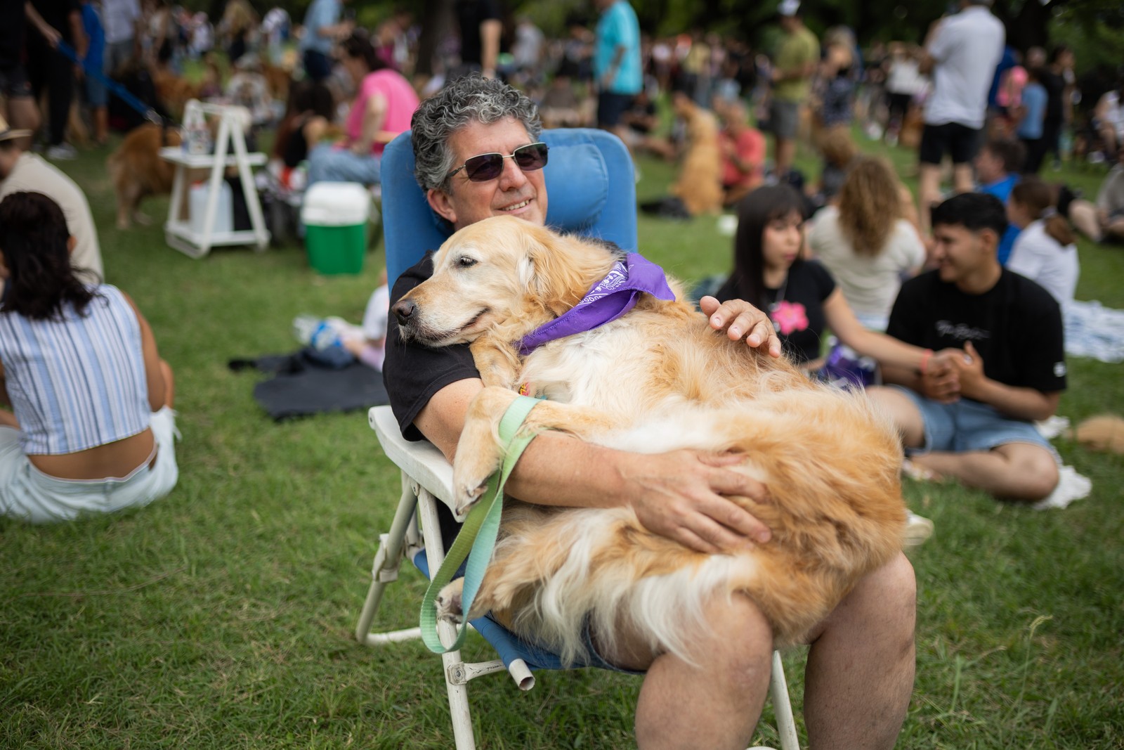 A golden retriever owner sits with his dog on his lap in a park, surrounded by many other people.