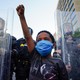 A young black boy wearing a mask raises his fist in front of a line of police officers using riot shields