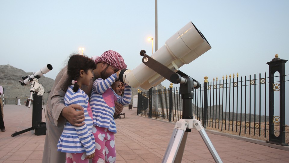 A father holds his two daughters while they look through a telescope.