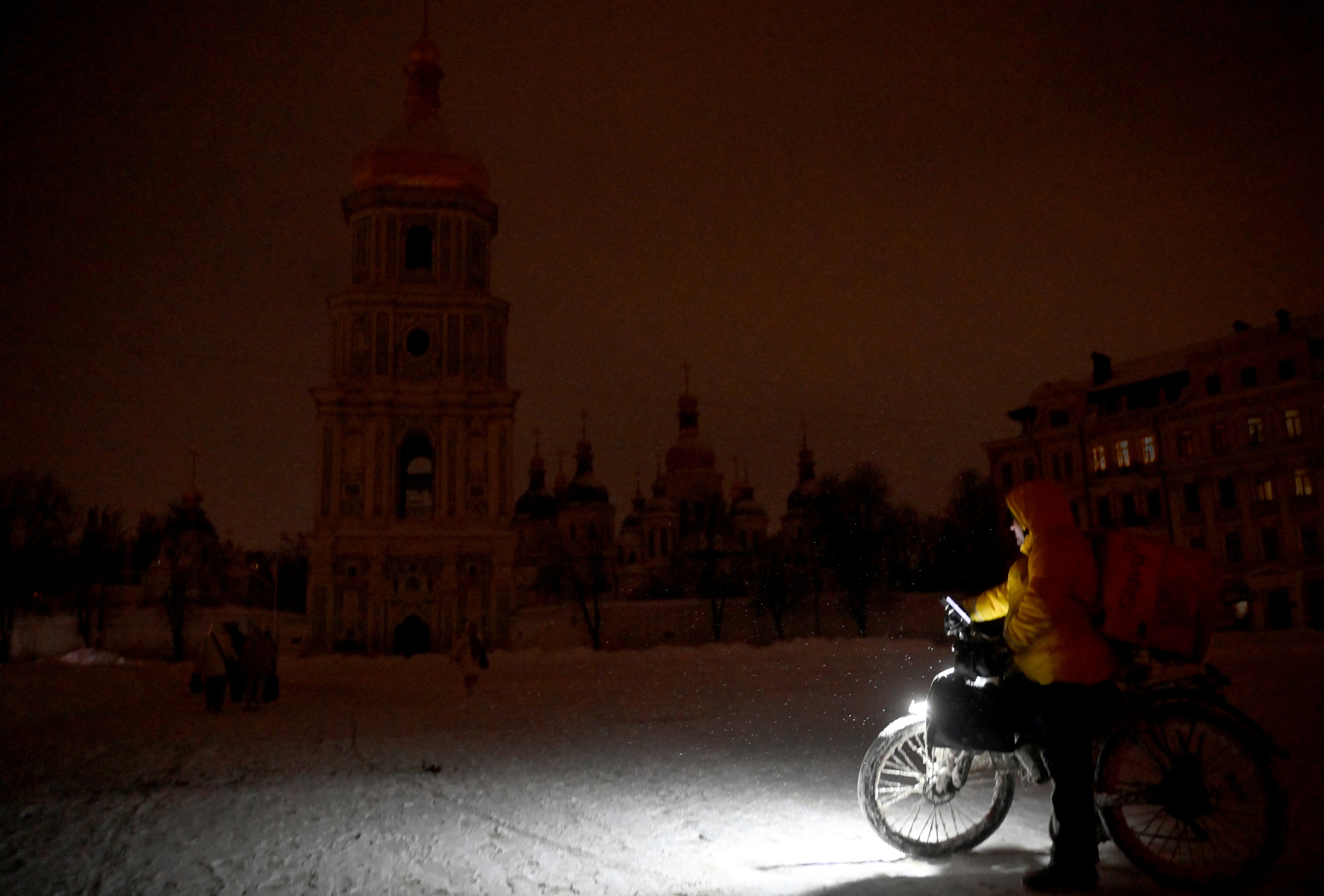 A delivery driver stands beside a motorcycle, with only its headlight lighting up the area, during a blackout.