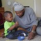 A father sits with his son on the carpet and looks at him lovingly while the child stares at a snack.