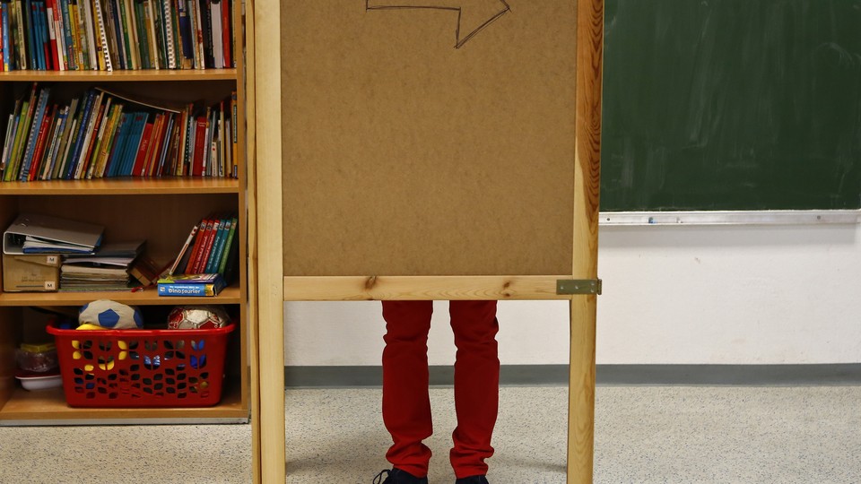 A pair of legs are visible behind a wooden corkboard in a classroom. 