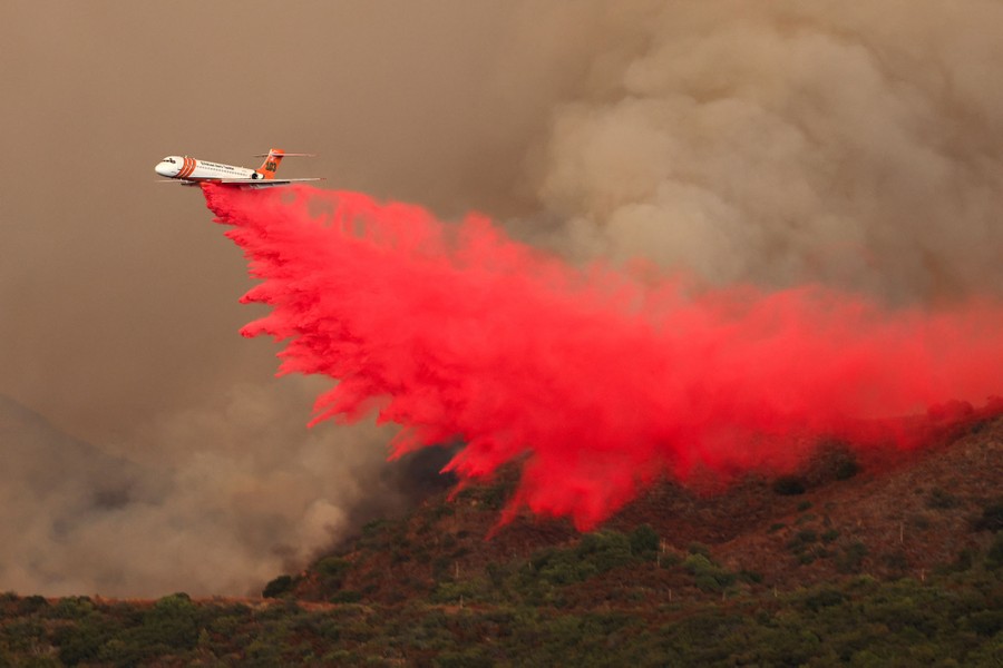 An aircraft drops a plume of bright-red fire retardant onto a hillside, with smoke rising in the background.