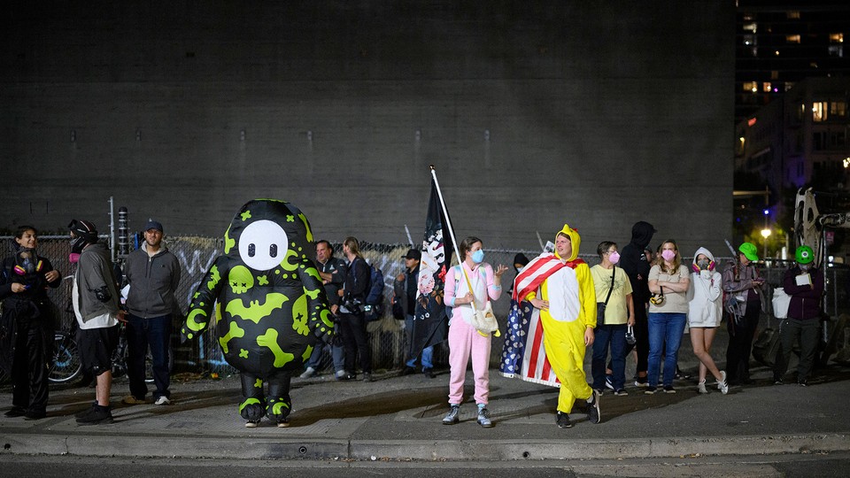 Protesters—including one dressed in a chicken suit—gather after dark outside a big gray building surrounded by a chain-link fence.