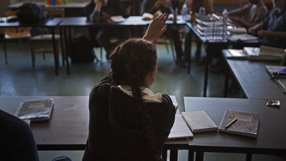 A student's back is photographed as she raises her hand. She wears her hair in long pigtail braids.