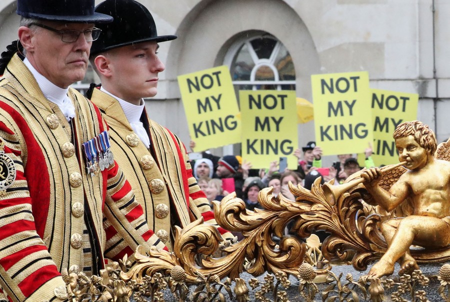 Protest signs reading "Not my King" are seen beyond a passing royal carriage.