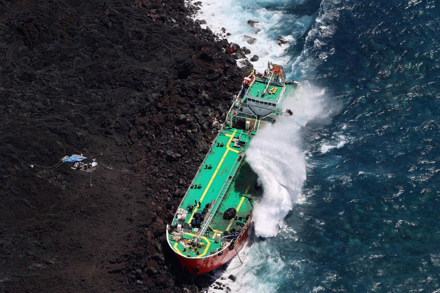 A large tanker ship lies stranded on a rocky shoreline, pounded by waves.